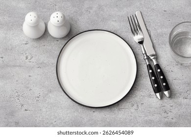 White empty plate with black rim, fork, knife, salt shakers, and glass on a gray textured surface. Minimalist dining concept, perfect for food-related visuals. - Powered by Shutterstock - Get 15% off with code: PIKWIZARD15