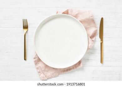 White Empty Plate And Golden Cutlery With Pink Napkin On White Wooden Table. Top View On Table Setting.