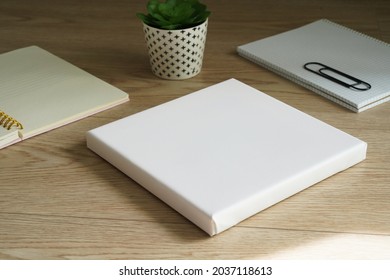 White Empty Square Canvas For Painting Lies On A Wooden Table Next To Two Notes And A Small Flower. Mockup Canvas