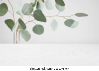 White Empty Table Mockup With Fresh, Green Eucalyptus Branches In The Background. Indoor Setting With Neutral Colours. For Displaying Digital Product Mockups. Suitable For Organic Or Natural Products.