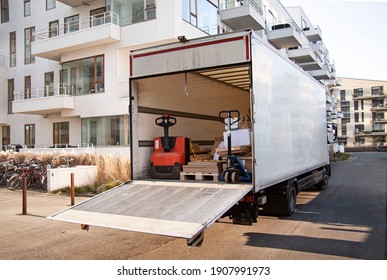 White Empty And Unidentified Delivery Truck With Tailgate Open In Front Of Modern Buildings. Forklifts And Packages On Pallets Inside The Storage Space Of The Lorry.