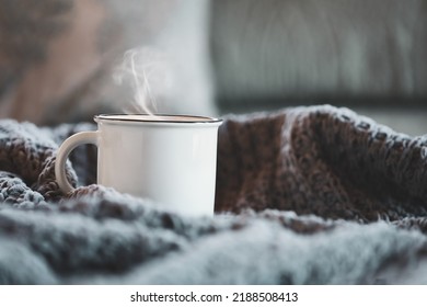 White enamel cup of hot steaming coffee sitting on a cozy bed surrounded by throw pillows and knit blankets. Selective focus on mug with extreme blurred foreground and background. - Powered by Shutterstock