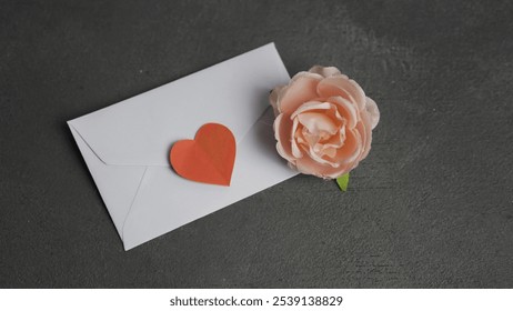 A white envelope with a heart on it sits on a grey surface. A pink flower is placed on top of the envelope - Powered by Shutterstock