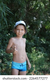 A white European schoolboy in swimming trunks and a hat shows a thumbs-up gesture against the backdrop of tropical foliage. Cool summer vacation. Copy space - Shutterstock ID 2264577277