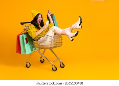 White excited woman laughing and using cellphone in shopping cart isolated over yellow background