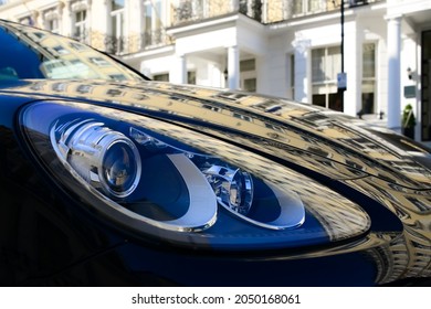 White facade of a typical old London town building reflected on the bonnet or engine hood of dark blue sports car parking in front of the renovated house. Distorted warped mirror image on varnish