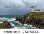 White Fanad Head Lighthouse contrasts against stormy skies and vivid blue ocean waters, with waves crashing against the rocky shore