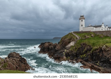 White Fanad Head Lighthouse contrasts against stormy skies and vivid blue ocean waters, with waves crashing against the rocky shore - Powered by Shutterstock