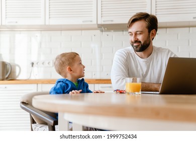 White father and son talking and using laptop while sitting at table in kitchen