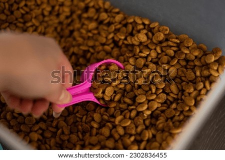 White female hand scooping up a portion of brown dog food kibble with a small bright pink measuring cup spoon from a plastic container to feed pet toy poodles