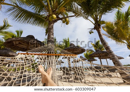 White female legs on hammock on tropical beach with palm leaf thatch roofing umbrellas and palm trees in the background