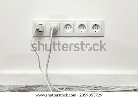 White five-way wall power socket installed on the white wall with inserted phone adapter and a white electrical plug, front view.