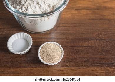 White Flour In A Glass Bowl Container, Dry Yeast, And Salt In Small Ramekin On Wood Table Surface.