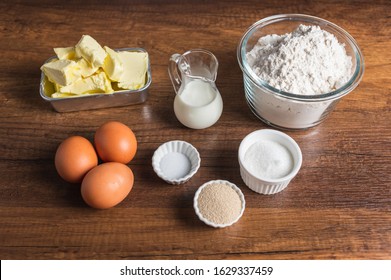 White Flour In A Glass Bowl Container, Dry Yeast, Salt, And Sugar In Small Ramekin. 3 Pieces Of Brown Chicken Eggs, Butter, And Milk In Small Glass Jar On Wood Table Surface.