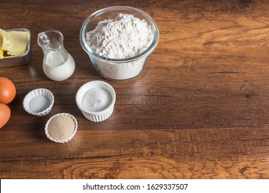 White Flour In A Glass Bowl Container, Dry Yeast, Salt, And Sugar In Small Ramekin. 3 Pieces Of Brown Chicken Eggs, Butter, And Milk In Small Glass Jar On Wood Table Surface.