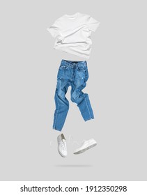 Weißes, fliegendes T-Shirt aus Baumwolle, blaue Jeans, weiße Turnschuhe aus Leder einzeln auf grauem Hintergrund. Sauberes weißes Unisex T-Shirt. Branding-Kleidung. Mock nach deinem Design. Sommerkleidung der Frauen im Frühling