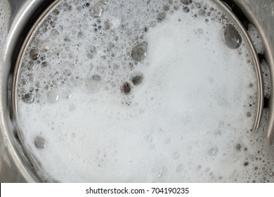 White foam with bubbles of clean in a washbasin. Drain hole with soap bubbles in metal sink, macro view. Mechanically adjustable drain plug closeup.