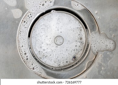 White foam with bubbles of cleaner in a washbasin. Drain hole with soap bubbles in metal sink, macro view. Mechanically adjustable drain plug closeup.
