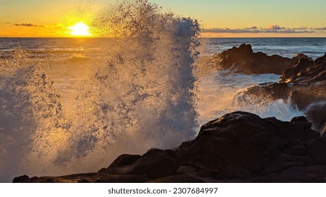 White foaming ocean waves crash up on a rocky cliff and sunset on the Pacific Ocean. Sun peaks behind wave as it sets with orange streaks contrasting blue ocean and cliffs extending into water - Powered by Shutterstock