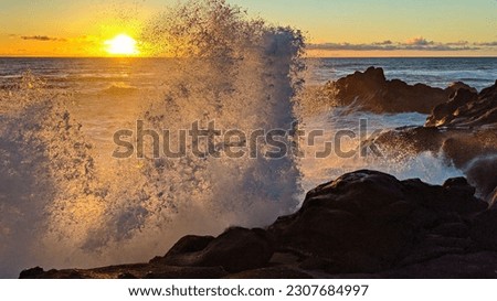 White foaming ocean waves crash up on a rocky cliff and sunset on the Pacific Ocean. Sun peaks behind wave as it sets with orange streaks contrasting blue ocean and cliffs extending into water