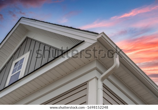 White frame gutter guard system, with gray horizontal\
and vertical vinyl siding fascia, drip edge, soffit, on a pitched\
roof attic at a luxury American single family home dramatic sunset\
sky
