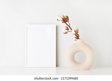 White Frame Mockup And Round Ceramic Vase With A Cotton Branch On A White Table By The Light Wall. Copy Space For Text