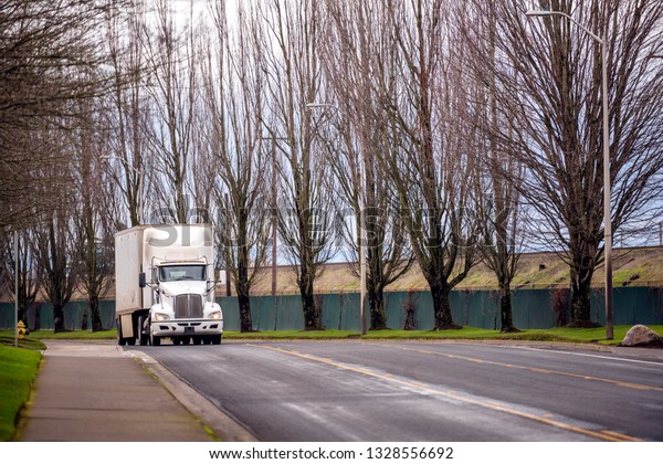 White freight transportation forward in American\
logistic industry big rig semi truck with compact heavy-duty box\
semi trailer moving on local road on city street with high bare\
trees on the side