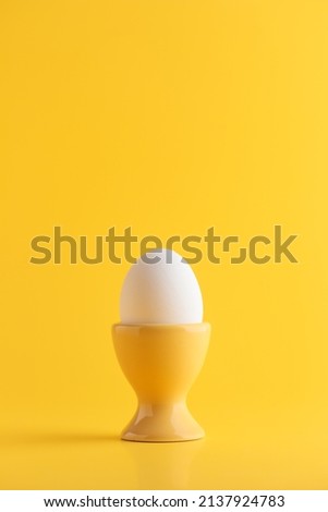 White fresh egg in the yellow egg stand, cup for breakfast on solid yellow background, vertical with empty space, Easter holiday, healthy eating