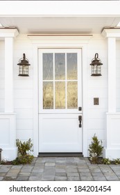 A White Front Door To A Classic White Family Home In Daytime. Door Features Intricate Etched Detail. Also Seen Is Columns, A Stone Porch, And Light Fixtures. 