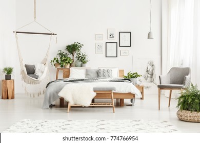 White fur on bench and grey chair in bohemian bedroom interior with hammock, plants and posters