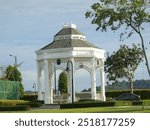 A white gazebo with intricate latticework stands in a park, surrounded by lush greenery and a tennis court in the background. The gazebo has a hexagonal shape and features a domed roof with decorative