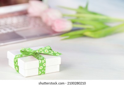 White gift box with a bright green ribbon stands on the table next to tulips and a laptop laptop. A bouquet of pink tulips is lying on the keyboard. the concept of online shopping for spring gifts
