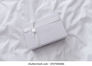 White Gift Box With Ribbon On White Wrinkled Bed Sheets Background. Minimalism Beauty Still Life, Wedding Present Concept. Copy Space, Mockup, From Above