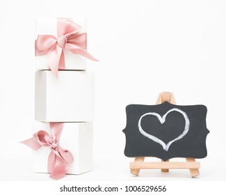 White Gift Boxes Of The Famous Jewelry Brand With Bracelets And Charms With A Pink Ribbon. Mock Up. Empty Place For An Inscription. Postcard For The Holiday. Heart Drawn Chalk On A Blackboard.