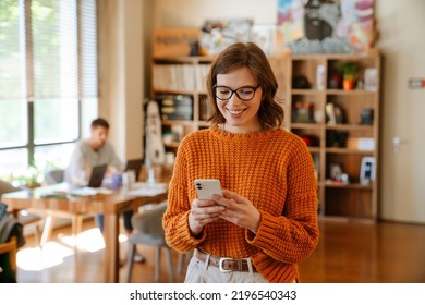 White ginger woman in eyeglasses using cellphone while working at office indoors - Shutterstock ID 2196540343