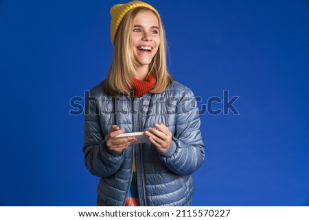 White girl wearing hat smiling and playing online game on cellphone isolated over blue background