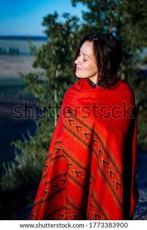 a white girl wrapped in a blanket enjoys life in the morning sun, orange plaid with an ethnic pattern, curly hair, peace and nature, against the background of the forest