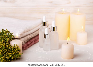 White glass dropper bottles with gel or lotion, oil or toner among burning candles. Towels and plant complete concept of spa treatments. Care and rest, beauty and health. Selective focus