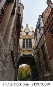 White and gold bridge linking two important buildings in Bruges, with three golden statues above, on a cloudy day.
