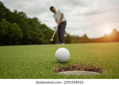 White golf ball rolling down golf hole on putting green with evening golf course backdrop and blurred golfers celebrating in winning tournament. - Powered by Shutterstock