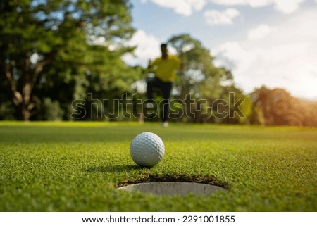 White golf ball rolling down golf hole on putting green with evening golf course backdrop and blurred golfers celebrating in winning tournament.