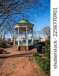 A white and green wooden gazebo on a red brick footpath surrounded by bare winter trees and lush green plants with a gorgeous clear blue sky at the Marietta Square in Marietta Georgia USA