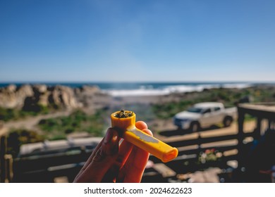 White hand holding a Yellow pipe with Marijuana (cannabis) in front of the ocean and the beach with rocks