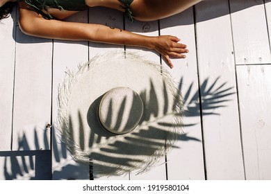 White hat on white wooden planks by the pool under the shade of a palm branch. A woman's body in a swimsuit is slightly visible