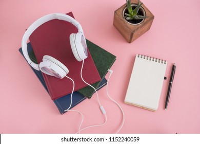 white headphones lie on pile of books on pink background. concept: audiobook