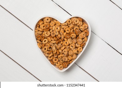 White Heart Shaped Bowl Of Cinnamon Flavored O Shaped Cereal And Oats On A White Wooden Background