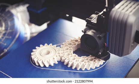 White Helical Gears Produced By Small Black FDM-printer On Blue Build Plate. Clean Bright Technical Scenery With Filament Rolls In Background. Excessive Selective Focus. Additive Manufacturing Concept