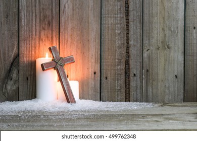 White holiday candles glowing behind wooden cross in snow by antique rustic wood background; Christmas, Easter and religious background with copy space