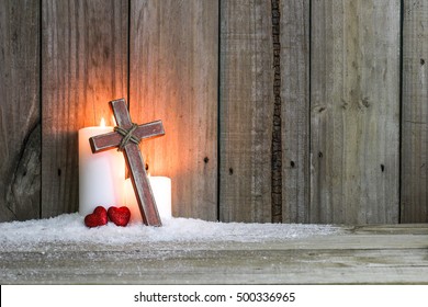 White Holiday Candles Glowing Behind Wooden Cross With Red Hearts In Snow By Antique Rustic Wood Background; Christmas, Easter And Religious Background With Copy Space