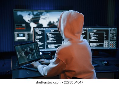 In white hoodie. Young professional female hacker is indoors by computer with lot of information on displays.
